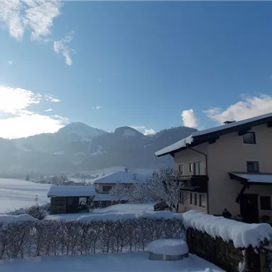 A winter landscape with snow-covered houses and mountains in the background. The sky is blue with a few clouds.