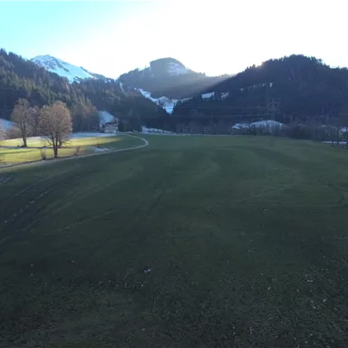 A wide green meadow surrounded by gentle hills and snow-capped mountains. The sun shines through the trees, creating a calm atmosphere.