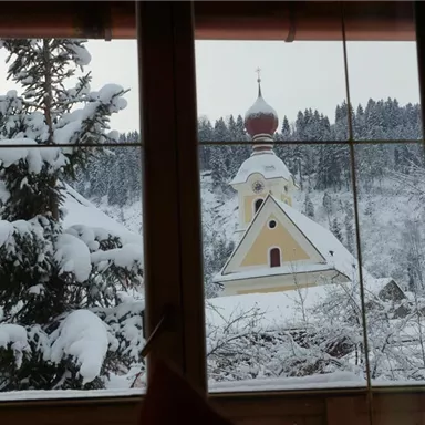 Eine verschneite Winterlandschaft mit einer Kirche im Hintergrund. Im Vordergrund sind schneebedeckte Bäume zu sehen.