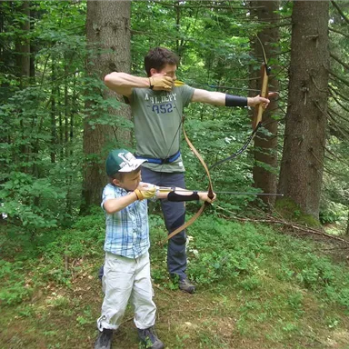 Ein Erwachsener und ein Kind üben das Bogenschießen im Wald. Sie stehen in einer grünen Umgebung zwischen hohen Bäumen.