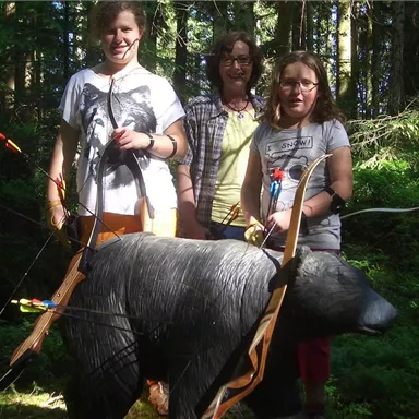 Drei Personen stehen im Wald neben einer bearbeiteten Bärenfigur mit Pfeilen. Es ist eine aktive Umgebung mit viel Grün und Sonnenlicht.