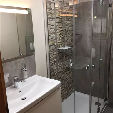 Ein modernes Badezimmer mit einer gläsernen Dusche und einem stilvollen Waschbecken. Die Wände sind mit natürlichem Stein verkleidet und die Beleuchtung wirkt warm und einladend.