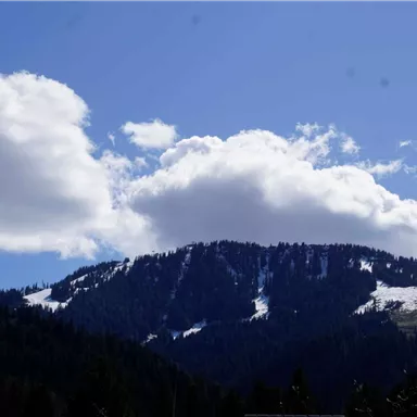 A picturesque mountain with snow-capped peaks and a blanket of clouds against a clear blue sky. The surroundings are surrounded by dense forests.