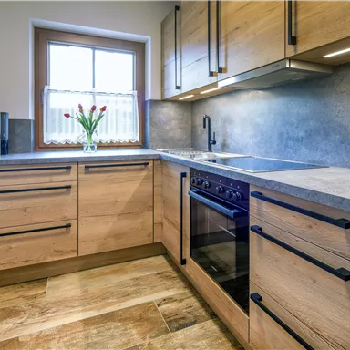 A modern kitchen with wooden cabinets and a gray work area. Fresh flowers in a vase are on the windowsill.