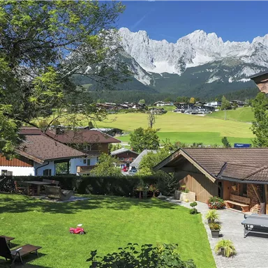 A picturesque garden with loungers, surrounded by traditional houses and majestic mountains in the background. The sky is clear and brilliantly blue.