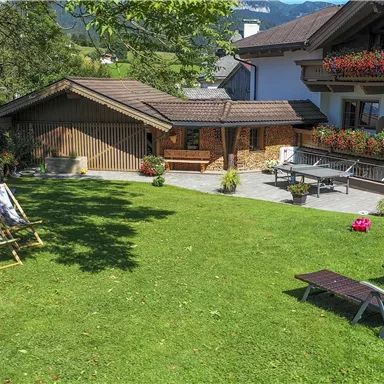 A quiet garden with loungers and artworks. In the background, a cozy wooden house and a two-story facade can be seen.