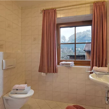 A cozy bathroom with a window that looks out at the mountains. The walls are tiled in a light color and the curtains are pink.