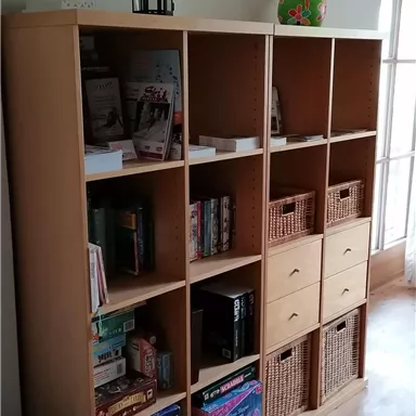 Ein Holzregal mit offenen und geschlossenen Fächern steht an der Wand. Darin sind Spiele, Bücher und verschiedene Korbwaren ordentlich verstaut.