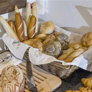 Ein Korb mit verschiedenen Brotsorten, darunter Baguettes, Brötchen und Croissants. Die Brote sind sorgfältig angeordnet und mit einem weißen Tuch bedeckt.