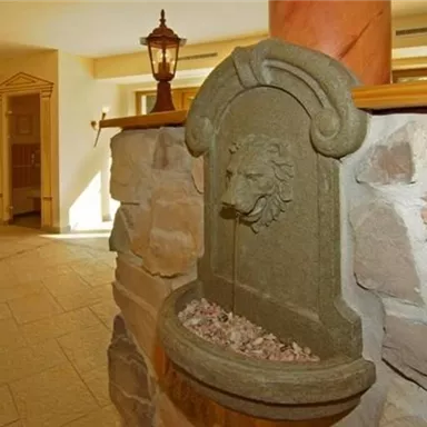 A decorative stone wall fountain with a lion motif. The fountain is located in a bright indoor space with natural stone elements.