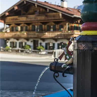 Ein malerischer Brunnen fließt mit klarem Wasser vor einem traditionellen, rustikalen Gebäude. Im Hintergrund sind Balkone mit Blumen zu sehen, die die Szene verschönern.