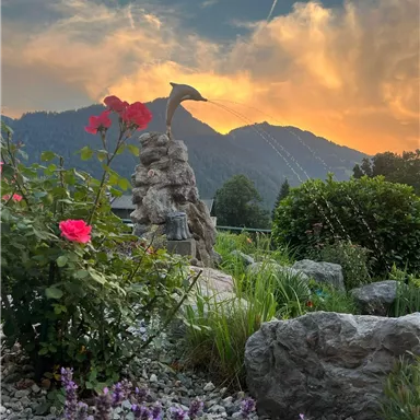 Ein schöner Garten mit roten Rosen und lila Blumen. Im Hintergrund erheben sich majestätische Berge unter einem farbenfrohen Himmel.