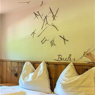 Ein gemütliches Schlafzimmer mit zwei Kissen und Holzvertäfelung. An der Wand sind kunstvolle Äste und der Schriftzug „Buchs“ zu sehen.