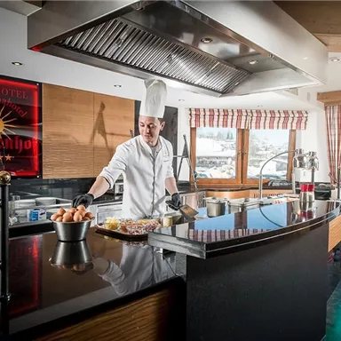 Ein Koch bereitet Essen in einer modernen Küche mit großen Fenstern und Blick auf die Berge vor. Die Atmosphäre ist einladend und gut ausgestattet.