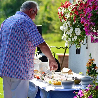 Ein Mann steht an einem Tisch mit verschiedenen Speisen im Freien. Im Hintergrund sind bunte Blumen und eine sonnige Umgebung zu sehen.