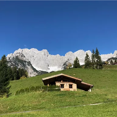 A cozy wooden house lies on a green meadow. In the background, impressive mountains and a blue sky can be seen.
