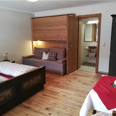 A cozy room with two beds and a sofa. The floor is made of wood and there is a small table with a decorative tablecloth.