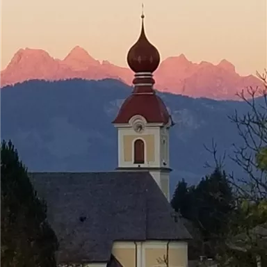 Eine Kirche mit einer markanten Kuppel und einem roten Dach steht vor majestätischen Bergen. Der Himmel ist in sanften Farben beleuchtet.