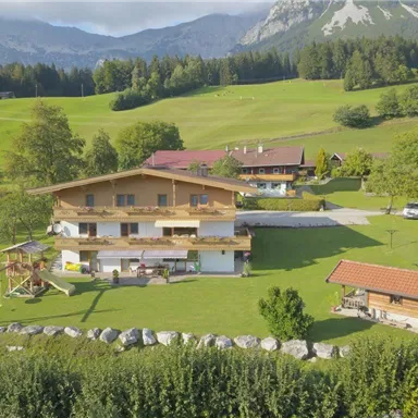 A picturesque landscape featuring several buildings and green meadows. In the background, there are mountains and a clear sky.