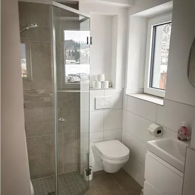 Ein modernes Badezimmer mit einer Glasdusche und einem weißen WC. Es gibt ein Fenster, das Licht hereinlässt, und einen kleinen Waschbecken-Unterschrank.