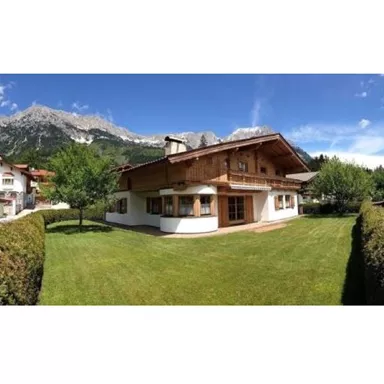 Ein charmantes Holzhaus mit einem gepflegten Garten und grünen Bäumen. Im Hintergrund erheben sich majestätische Berge unter einem klaren Himmel.