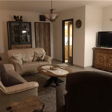 Ein gemütliches Wohnzimmer mit einem beigen Sofa und einem braunen Sessel. Es gibt einen Couchtisch und einen Fernseher, umgeben von rustikalen Möbeln.