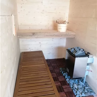 Ein gemütlicher Raum mit Holzwänden, einer Holzbank und einem Saunaofen. Auf dem Regal oben steht ein Holzbehälter.