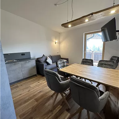 Ein modernes Wohnzimmer mit einer Küchenzeile und einem großen Esstisch. Die Möbel sind gemütlich und der Raum hat viel Tageslicht.