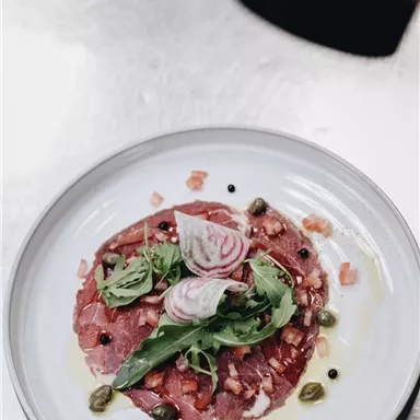 Ein kunstvoll angerichteter Teller mit Carpaccio, garniert mit Rucola, Zwiebeln und Kapern. Die Farben und Texturen machen das Gericht ansprechend und appetitlich.