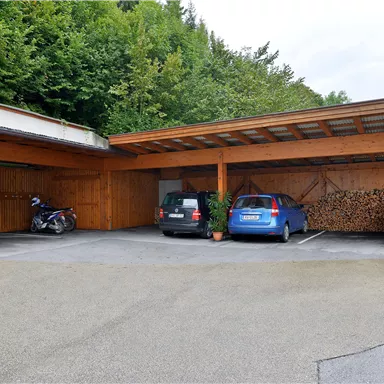 Ein überdachter Parkplatz mit zwei Autos und einem Motorrad. Im Hintergrund sind Holzstapel und viel Grün zu sehen.