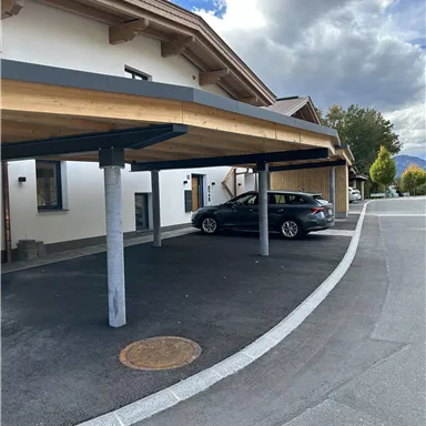 Eine moderne Überdachung für Autos vor einem Gebäude. Im Hintergrund sind Wolken und Bäume zu sehen.