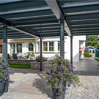 Eine überdachte Terrasse mit modernen Möbeln und blühenden Pflanzen. Im Hintergrund sind ein Haus und ein kleiner Garten sichtbar.