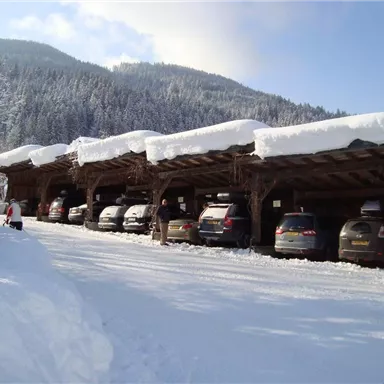 A parking lot with covered parking spaces, surrounded by snow and mountains. In the background, people can be seen walking in the snow.