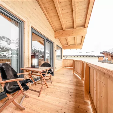 Ein gemütlicher Balkon mit Holzboden und zwei Stühlen. Im Hintergrund sind schneebedeckte Berge und eine ruhige Landschaft zu sehen.