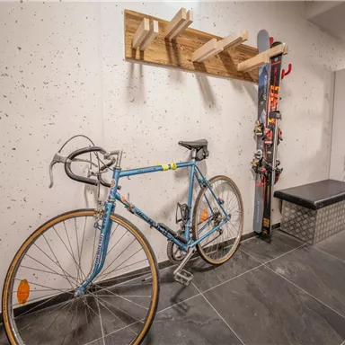 Ein Fahrrad steht in einem modernen Raum mit einer grauen Wand. Neben dem Fahrrad befindet sich eine Skiaufbewahrung mit Skiern und Stöcken.