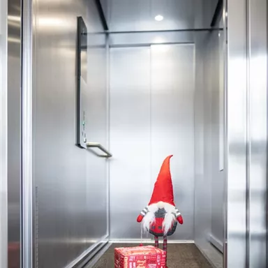 Ein Aufzug mit einem kleinen Weihnachtsmann und einem Geschenk. Die Wände sind aus Metall und das Licht ist hell.
