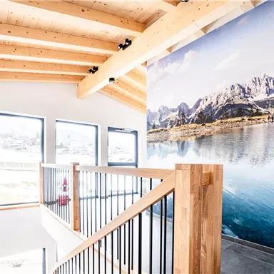 Ein moderner Treppenaufgang mit Holzgeländer. An der Wand ist ein schönes Landschaftsbild mit Bergen und einem klaren See zu sehen.
