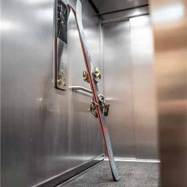 Ein Aufzug mit einer metallischen Wand. Im Vordergrund lehnen Skier an der Wand.
