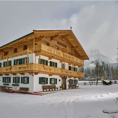 Ein schönes Holzhaus im Schnee, umgeben von einer winterlichen Landschaft. Die Fassade hat grüne Fensterläden und einen traditionellen alpenländischen Stil.