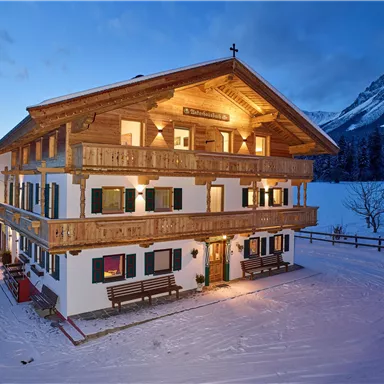 Ein charmantes Holzhaus im Winter mit beleuchteten Balkonen. Im Hintergrund sind schneebedeckte Berge und ein klarer Himmel zu sehen.