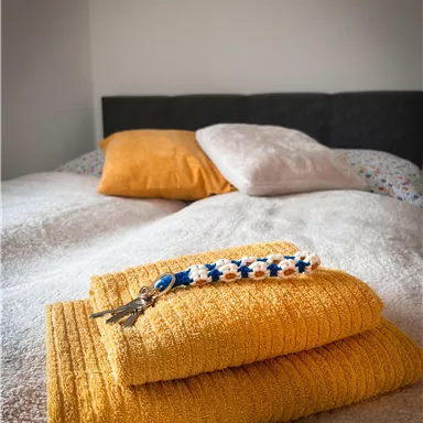 Ein gemütliches Bett mit orangefarbenen Kissen und frischen, gelben Handtüchern. Am Handtuch liegt ein farbiger Schlüsselanhänger.