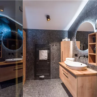 Ein modernes Badezimmer mit eleganten Holzmöbeln und einer großen Spiegelrunde. Die Wände sind mit dunkelgrauem Stein verkleidet, was dem Raum einen stilvollen Look verleiht.