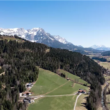 Eine wunderschöne Berglandschaft mit schneebedeckten Gipfeln und saftig grünen Wiesen. Im Vordergrund sind vereinzelte Gebäude und ein klarer Fluss zu sehen.