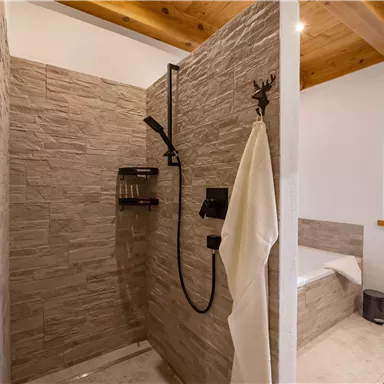 A modern shower with an elegant tiled wall and a stylish rain shower. Next to the shower is a freestanding bathtub and a window that lets in daylight.