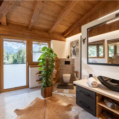 A modern bathroom with a wooden ceiling and large windows. There is an elegant washbasin, a shower, and a plant for a pleasant atmosphere.