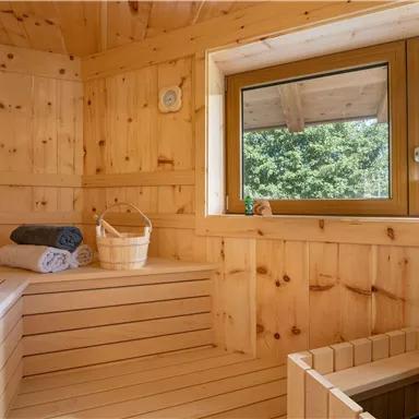 A cozy sauna room made of light wood with a window view into the greenery. On the bench are towels and a basket.