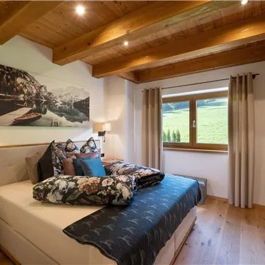 A cozy bedroom with wooden ceilings and a large window. The bed is decorated with colorful pillows and blankets, and a landscape painting hangs on the wall.