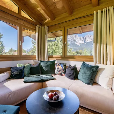A cozy living area with a large sofa and many cushions. Through the windows, one has a view of the mountains and nature.