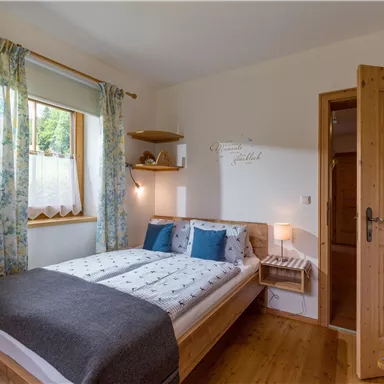 Ein gemütliches Schlafzimmer mit einem Doppelbett und blauen Kissen. Die Wände sind hell und durch das Fenster strömt Tageslicht herein.