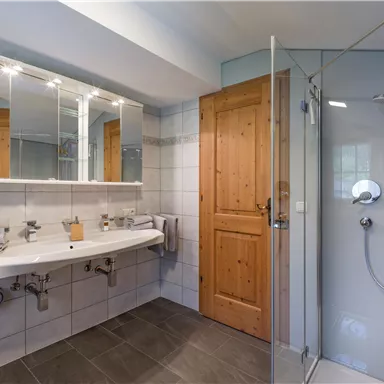 Ein modernes Badezimmer mit einer Dusche, einem Doppelwaschbecken und großen Spiegeln. Der Raum hat helle Fliesen und eine Holzhaustür.